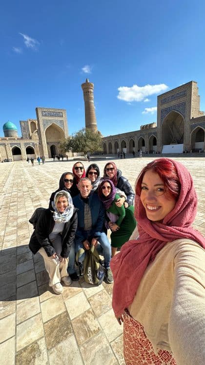 A woman takes a selfie of her WeRoad group trip, with women wearing headscarves, in a sunny courtyard with a historic minaret.