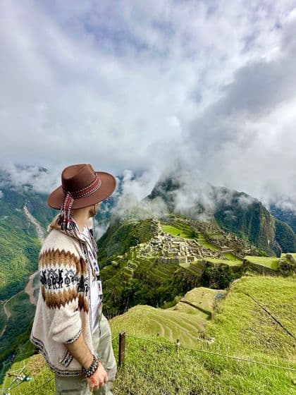 Un uomo con un cappello e un maglione a fantasia osserva antiche rovine costruite su verdi montagne terrazzate sotto un cielo nuvoloso.