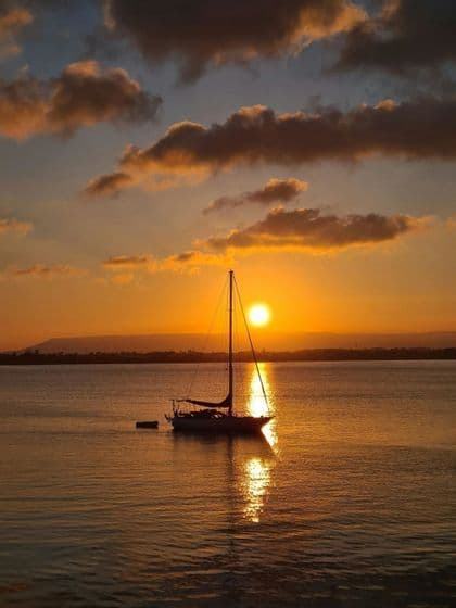 Una barca a vela è ancorata sull'acqua durante un tramonto dorato, con il suo riflesso che luccica sulla superficie.