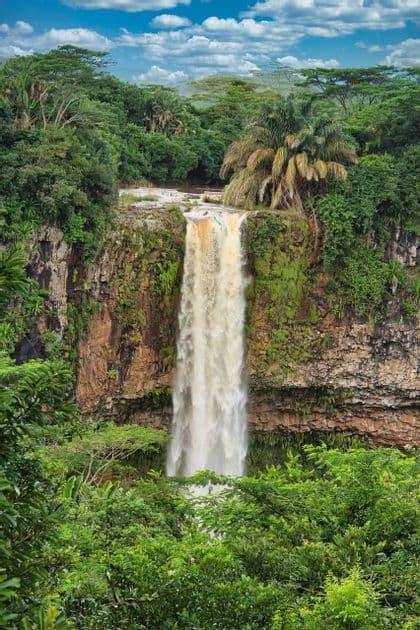Une cascade se jette d'une falaise moussue dans une jungle tropicale dense sous un ciel bleu parsemé de nuages blancs.
