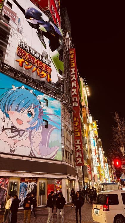 Alti edifici coperti da grandi cartelloni anime e insegne al neon illuminano una trafficata via della città giapponese di notte.