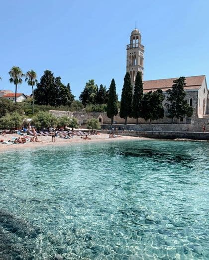 Gente tomando el sol en una playa de arena junto a agua turquesa cristalina, con una gran iglesia de piedra y un campanario al fondo.