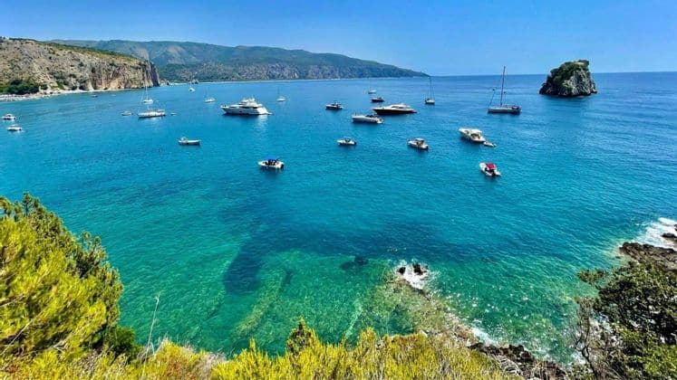 Veduta dall'alto di numerose imbarcazioni e yacht su acque limpide e turchesi in una baia costiera con scogliere sullo sfondo.