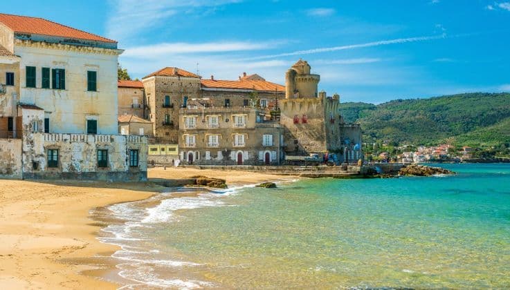 Una spiaggia di sabbia con acque cristalline turchesi di fronte a una cittadina costiera con una fortezza in pietra e colline verdi sullo sfondo.