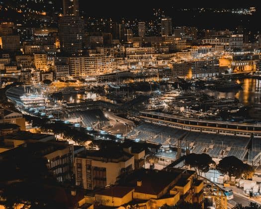 Un porto cittadino illuminato di notte, pieno di yacht e barche, con edifici e una tribuna che brillano sullo sfondo.