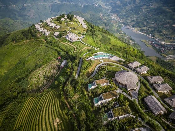 Vista aérea de un resort con villas y piscinas en una exuberante montaña verde, con vistas a campos en terrazas y un valle fluvial.