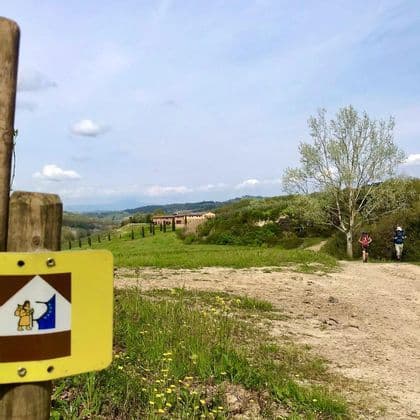 Due escursionisti con zaini seguono un sentiero sterrato attraverso un paesaggio verde e collinare, con un segnavia in primo piano.