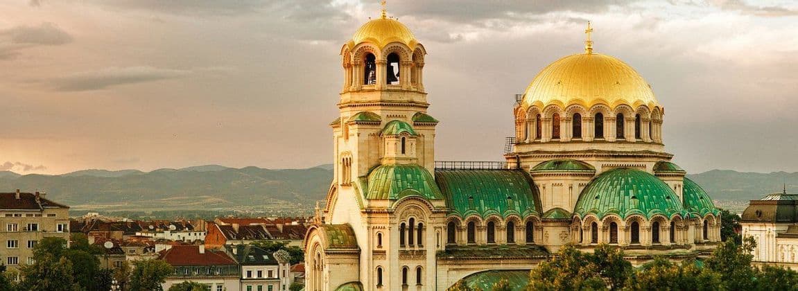 Una grande cattedrale con cupole dorate e tetti verdi si affaccia su una città con montagne lontane sotto un cielo nuvoloso.