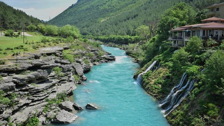 Un río turquesa vibrante serpentea a través de un cañón rocoso, con pequeñas cascadas que caen por una exuberante ladera verde junto a un edificio.