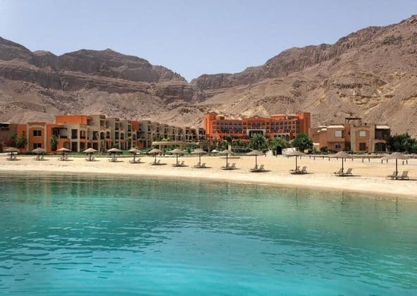 Un hotel resort en una playa de arena con tumbonas y sombrillas, frente a tranquilas aguas turquesas al pie de montañas áridas.