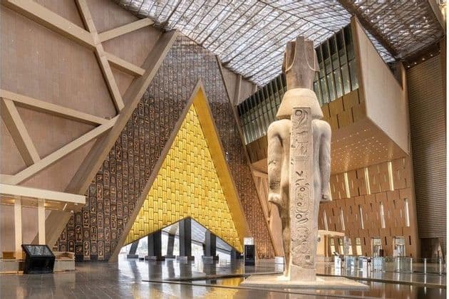 Una colosal estatua del antiguo Egipto se alza en una sala de museo moderna junto a una pared triangular decorada con oro y jeroglíficos.