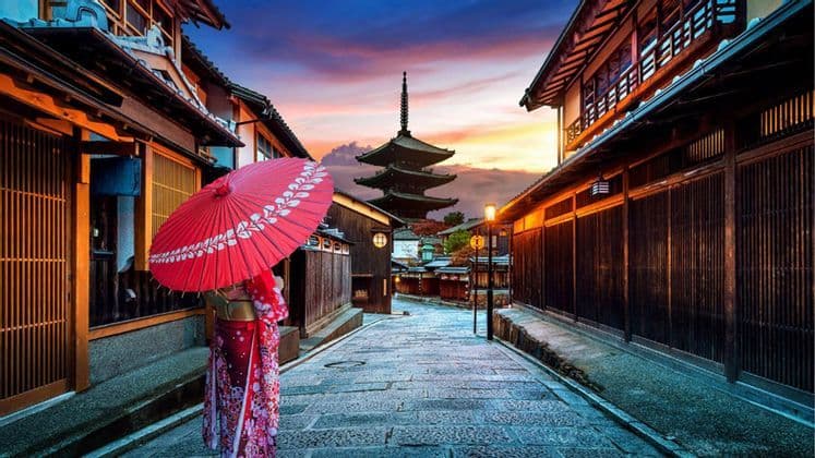 Una persona en kimono con un paraguas rojo en una calle de piedra de un pueblo tradicional, con una pagoda visible al atardecer.