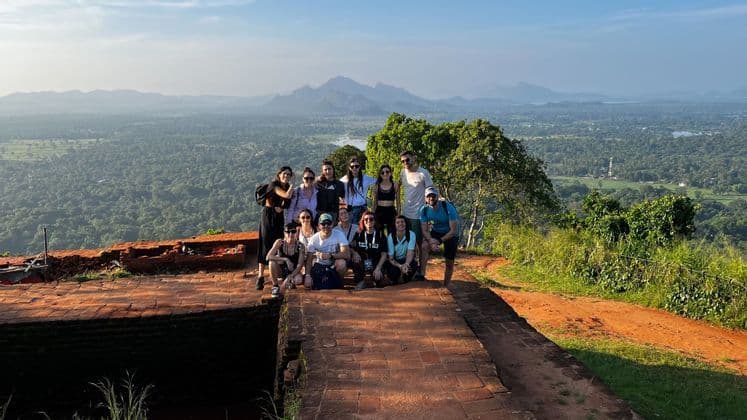 Un groupe WeRoad en voyage, posant pour une photo depuis un point de vue en brique, dominant une vaste vallée verdoyante et des montagnes lointaines.