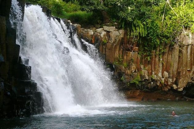 Eine Person schwimmt in einem Naturbecken am Fuße eines breiten Wasserfalls, der über dunkle, säulenartige Felsformationen stürzt.