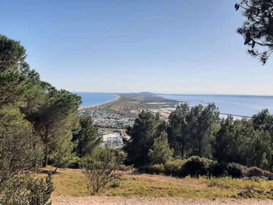 Une vue panoramique depuis une colline montrant une ville côtière et un long cordon littoral séparant la mer d'une lagune sous un ciel clair.