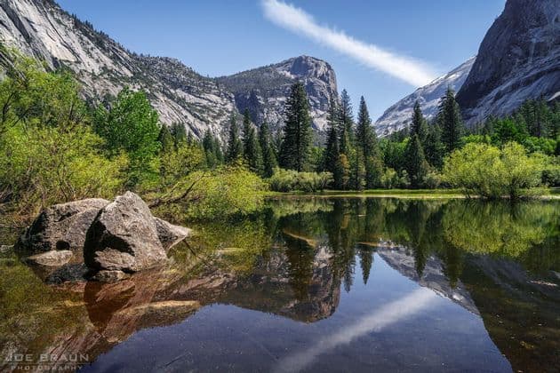 Un lago placido riflette montagne di granito, alberi verdeggianti e un cielo azzurro limpido.