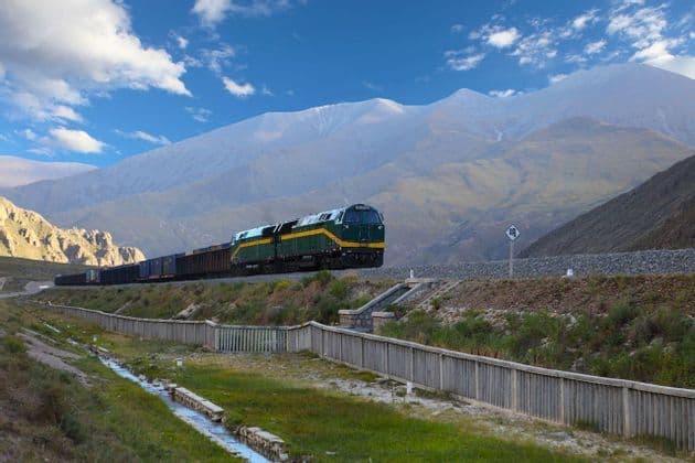 Un lungo treno merci verde e giallo viaggia sui binari attraverso una valle di montagna con un ruscello in primo piano.