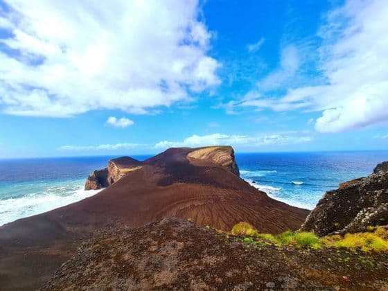 Un promontoire volcanique brun s'avance dans l'océan bleu, avec des vagues se brisant sur le rivage sous un ciel partiellement nuageux.
