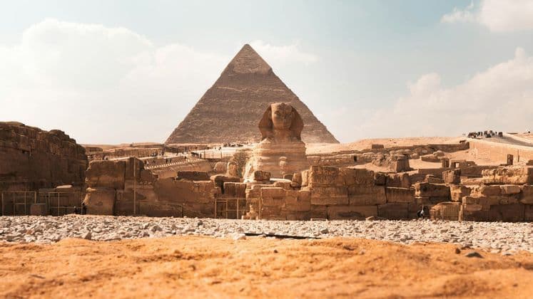 Una vista desde el suelo de la Gran Esfinge con una gran pirámide al fondo, rodeada de antiguas ruinas de piedra bajo un cielo brillante.