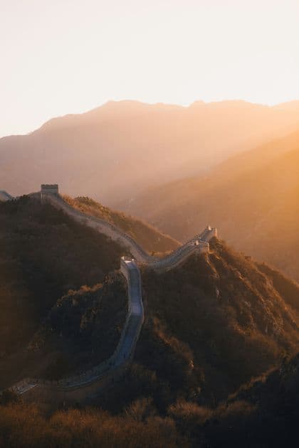 Una lunga muraglia di pietra con torri di guardia si snoda lungo una catena montuosa durante un tramonto dorato e velato.
