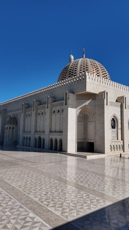Una mezquita de piedra ornamentada con una gran cúpula estampada y arcos, vista desde su patio embaldosado bajo un cielo azul claro.