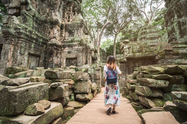 Una mujer con una mochila camina por un sendero de madera a través de antiguas ruinas de piedra cubiertas de musgo y rodeadas de árboles.