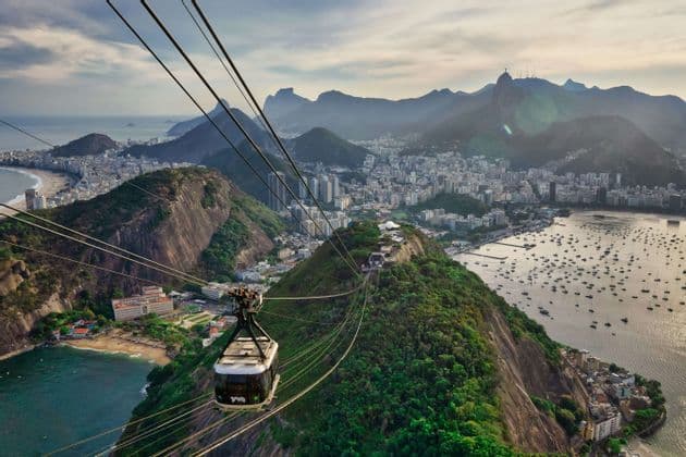 Un teleférico se desliza sobre una montaña exuberante, con una extensa ciudad costera, una bahía con barcos y montañas al fondo.