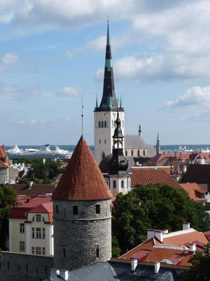 Eine Luftaufnahme einer historischen Stadt mit einem Steinturm mit rotem Dach, einem hohen Kirchturm und einem Kreuzfahrtschiff auf dem Wasser.