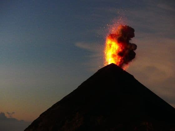 Un volcan en silhouette entre en éruption avec de la lave et de la fumée sur un ciel crépusculaire.