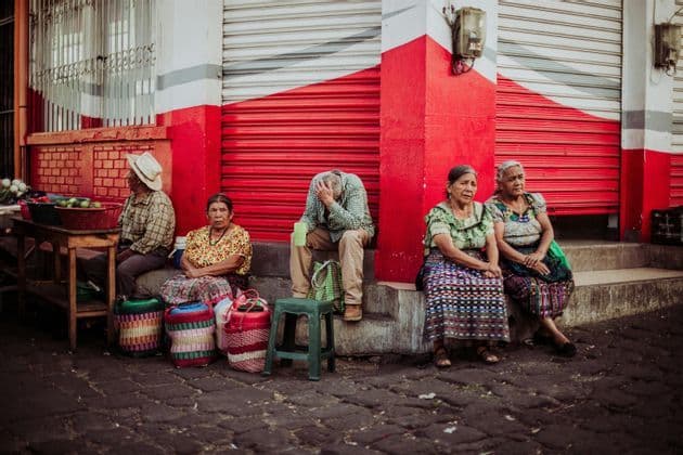 Cinq habitants, quatre femmes et un homme, se reposent sur les marches d'un bâtiment avec un volet rouge dans une rue pavée.