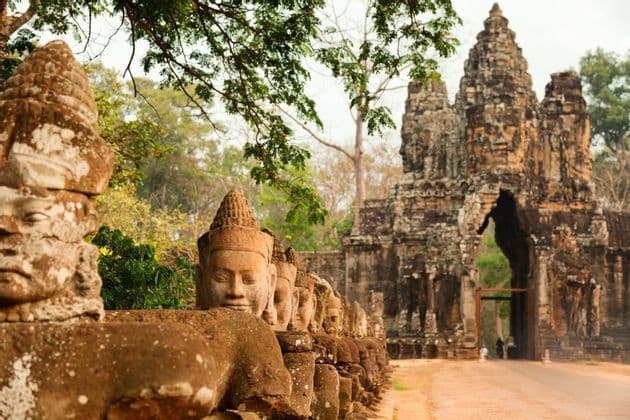A row of weathered stone statues with carved faces leads to an ancient, ornate stone gate under lush green trees.