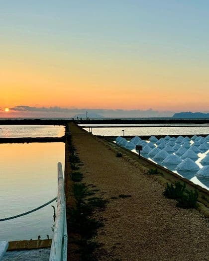 Un camino de grava separa las salinas con montículos de sal blanca bajo un cielo de atardecer sobre el mar.