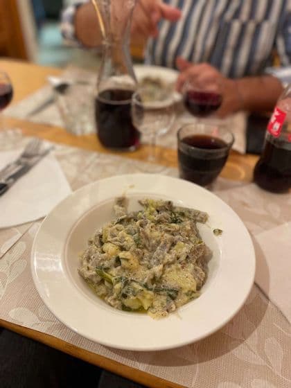 Primo piano di una ciotola bianca di pasta cremosa con verdure e patate, servita su un tavolo da pranzo con vino rosso.