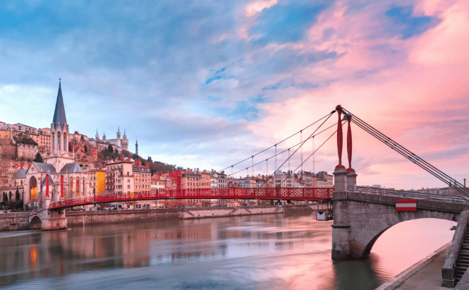 Un ponte rosso attraversa un fiume con una città collinare e una chiesa sullo sfondo sotto un cielo rosa e blu.