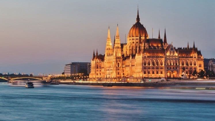 Un edificio gubernamental ornamentado iluminado con luces doradas al anochecer, visto desde el otro lado de un río con barcos.