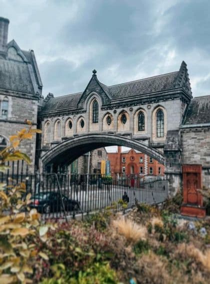 Un puente de piedra con arco de estilo gótico que conecta dos grandes edificios sobre una calle, visto desde detrás de una valla y un jardín.