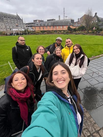 Un grupo de WeRoad se toma una selfie sonriente en un parque verde con edificios históricos de fondo.