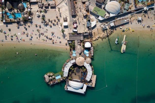 Una vista aerea di un beach club galleggiante con piscine che si estendono da una spiaggia sabbiosa in acqua turchese con barche.