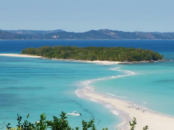 Eine sich schlängelnde weiße Sandbank verbindet sich mit einer kleinen, grünen Insel in türkisfarbenem Wasser, mit fernen Bergen im Hintergrund.