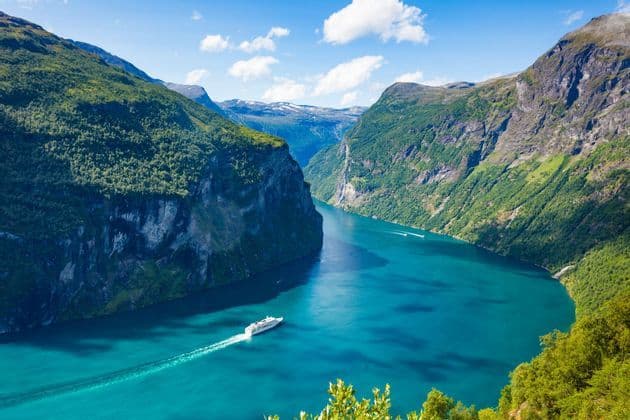 Vue aérienne d'un bateau de croisière blanc naviguant dans un fjord turquoise, entouré de montagnes vertes escarpées sous un ciel bleu.