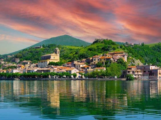Un borgo lacustre con una chiesa e case dai tetti in terracotta riflesse nell'acqua sotto un cielo al tramonto dai colori vivaci.