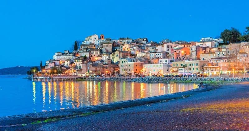 Un borgo collinare illuminato su un lungomare al tramonto, con le luci degli edifici che si riflettono sull'acqua calma.