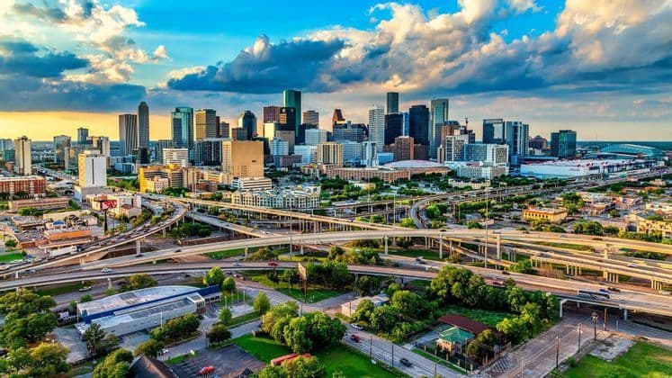 An aerial view of a modern city skyline with skyscrapers and highway interchanges under a blue and cloudy sky.