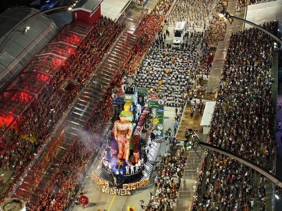 Una vista aerea di una grande parata di carnevale di notte, con un carro allegorico elaborato che si muove lungo una strada affollata di spettatori.