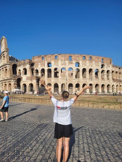 Ein WeRoad-Reiseleiter mit ausgestreckten Armen, von hinten gesehen, vor dem Kolosseum an einem sonnigen Tag.