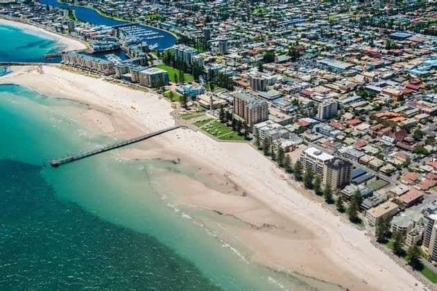 Eine Luftaufnahme einer Küstenstadt mit einem Sandstrand, einem langen Pier, der ins türkisfarbene Wasser ragt, und einem nahegelegenen Yachthafen.