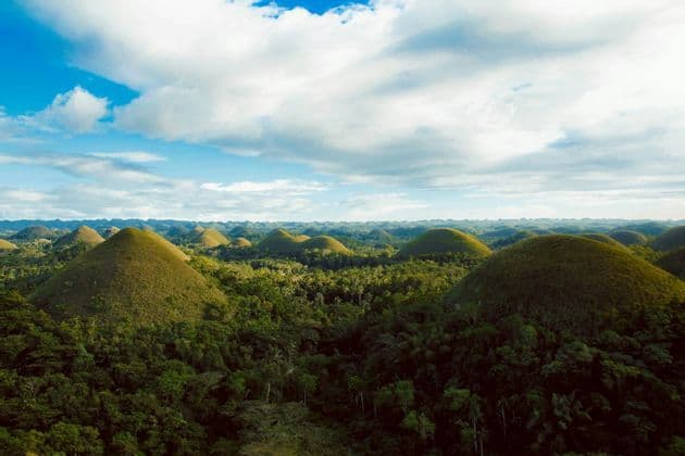 Una vista aérea de numerosas colinas verdes en forma de cono que se elevan desde un denso bosque bajo un cielo azul con nubes blancas.