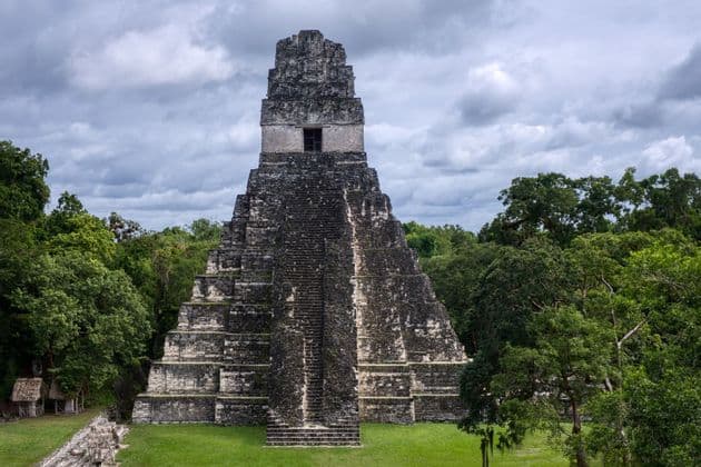 Una antigua pirámide escalonada de piedra emerge de una densa selva verde bajo un cielo nublado.