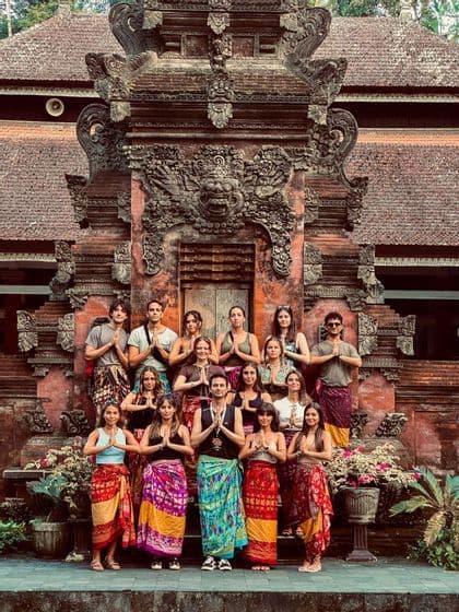 A WeRoad group trip wearing colorful sarongs poses with hands in a prayer gesture in front of an ornate stone temple.