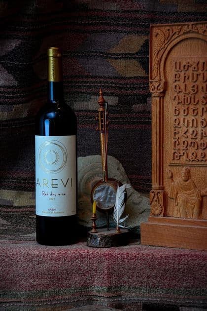 Una bottiglia di vino rosso armeno si trova accanto a uno strumento a corde tradizionale e una tavoletta di legno intagliato su un tessuto fantasia.